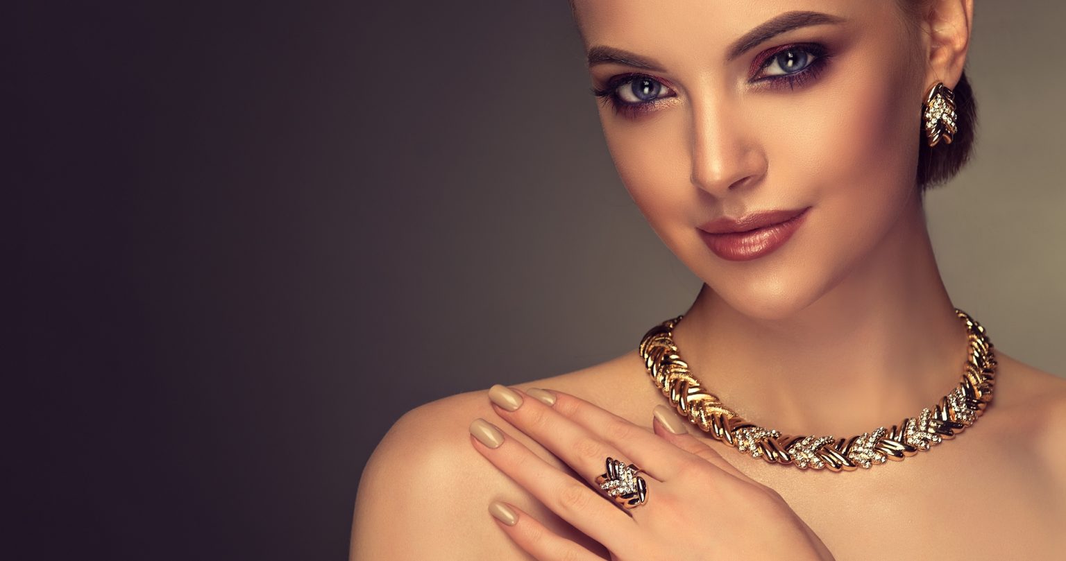 Portrait of gorgeous, young, woman illuminated by sophisticated multicolored light. Gilded jewelry set containing earrings, necklace and ring is dressed on young perfectly looking woman.